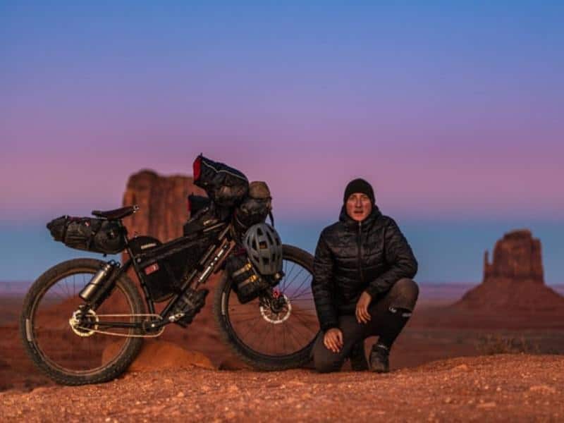 A man in black jacket and black beanie posing for a photo beside a loaded mountain bike