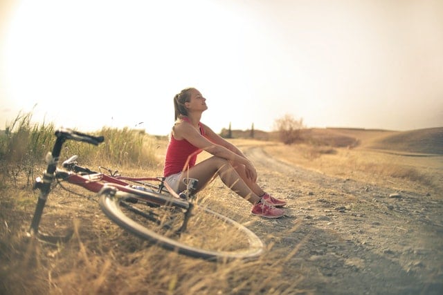 woman sitting on ground in unpaved road with her bike lying on a withered grass