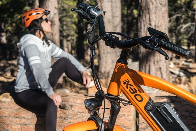 An image of orange e-bike parked in a place full of trees with the woman cyclist in the background