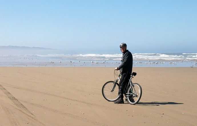 An elderly man standing with his bicycle, striking a pose for a photo against the backdrop of a serene beach.
