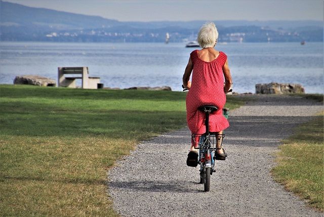 An elderly woman, dressed in a red duster dress, riding a bicycle toward a breathtaking sea view.